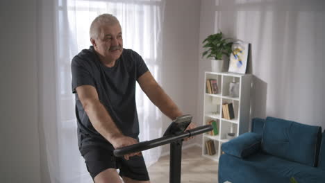 Ein-Müder-älterer-Mann-Dreht-Die-Pedale-Eines-Stationären-Fahrrads-Und-Trainiert-Allein-Zu-Hause,-Um-Körperliche-Aktivität-Und-Einen-Gesunden-Lebensstil-Beizubehalten